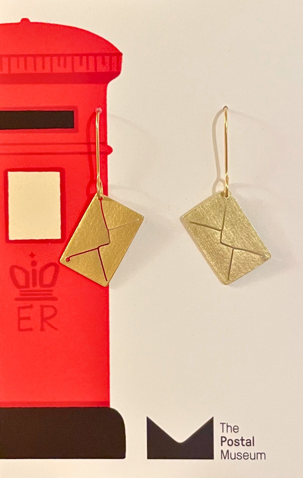 Gold envelope-shaped earrings in front of a red postbox with branding, on a beige background.