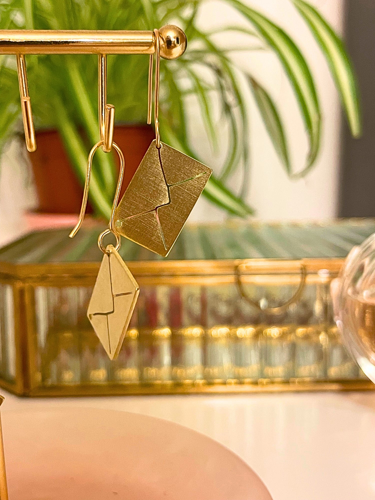 Gold earrings hanging on a gold stand with a blurred plant and decorative box in the background.