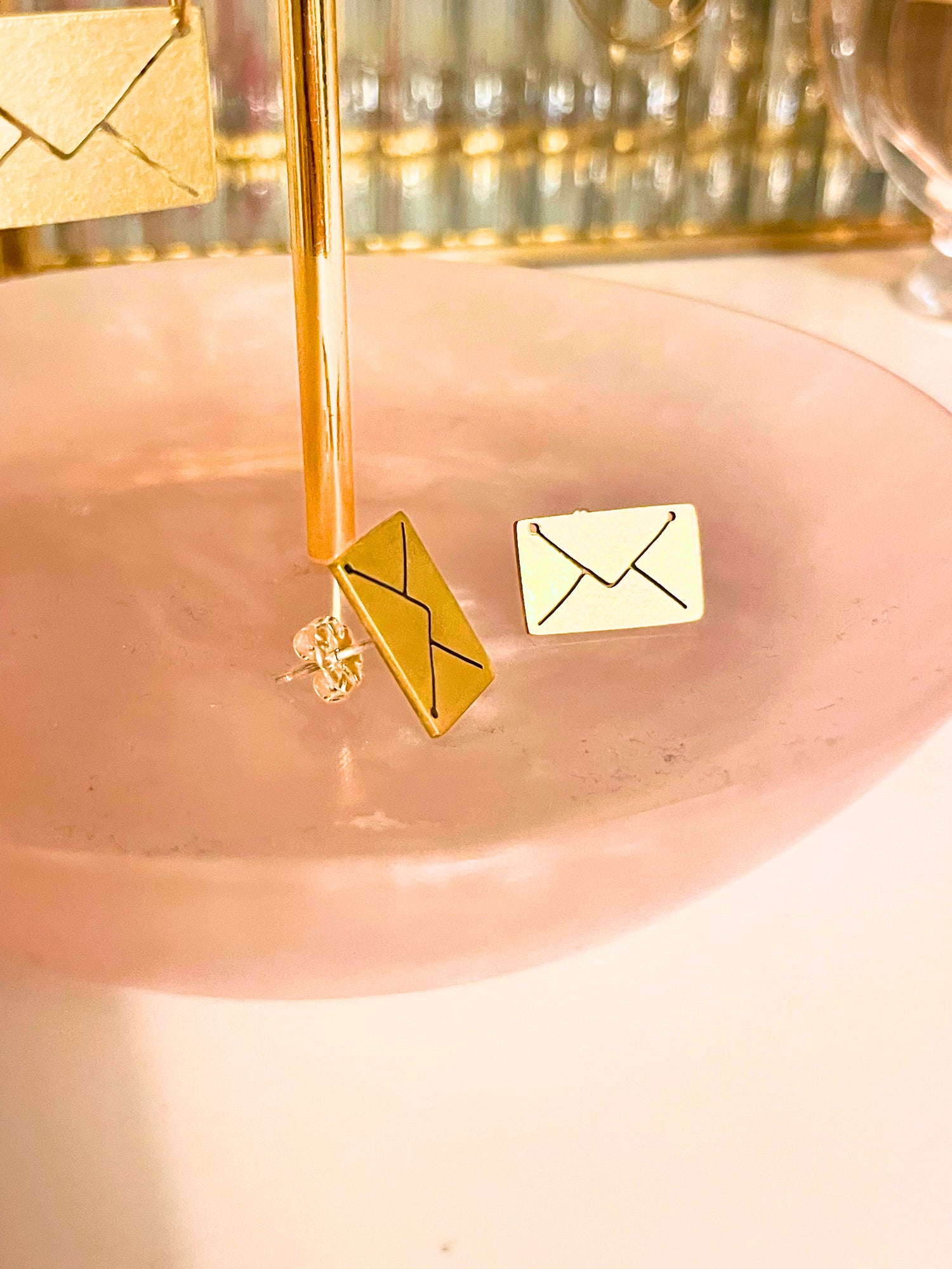 Gold envelope-shaped earrings on a pink surface with a blurred background